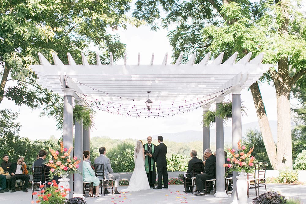 A bride and groom stand under a decorative pergola during their outdoor wedding ceremony, surrounded by guests.