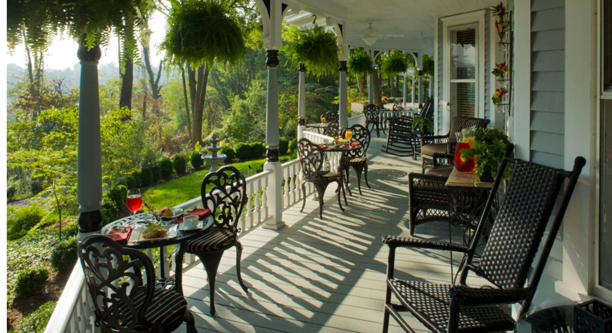 A charming porch with black wicker furniture, tables set for tea, and lush greenery in the background.