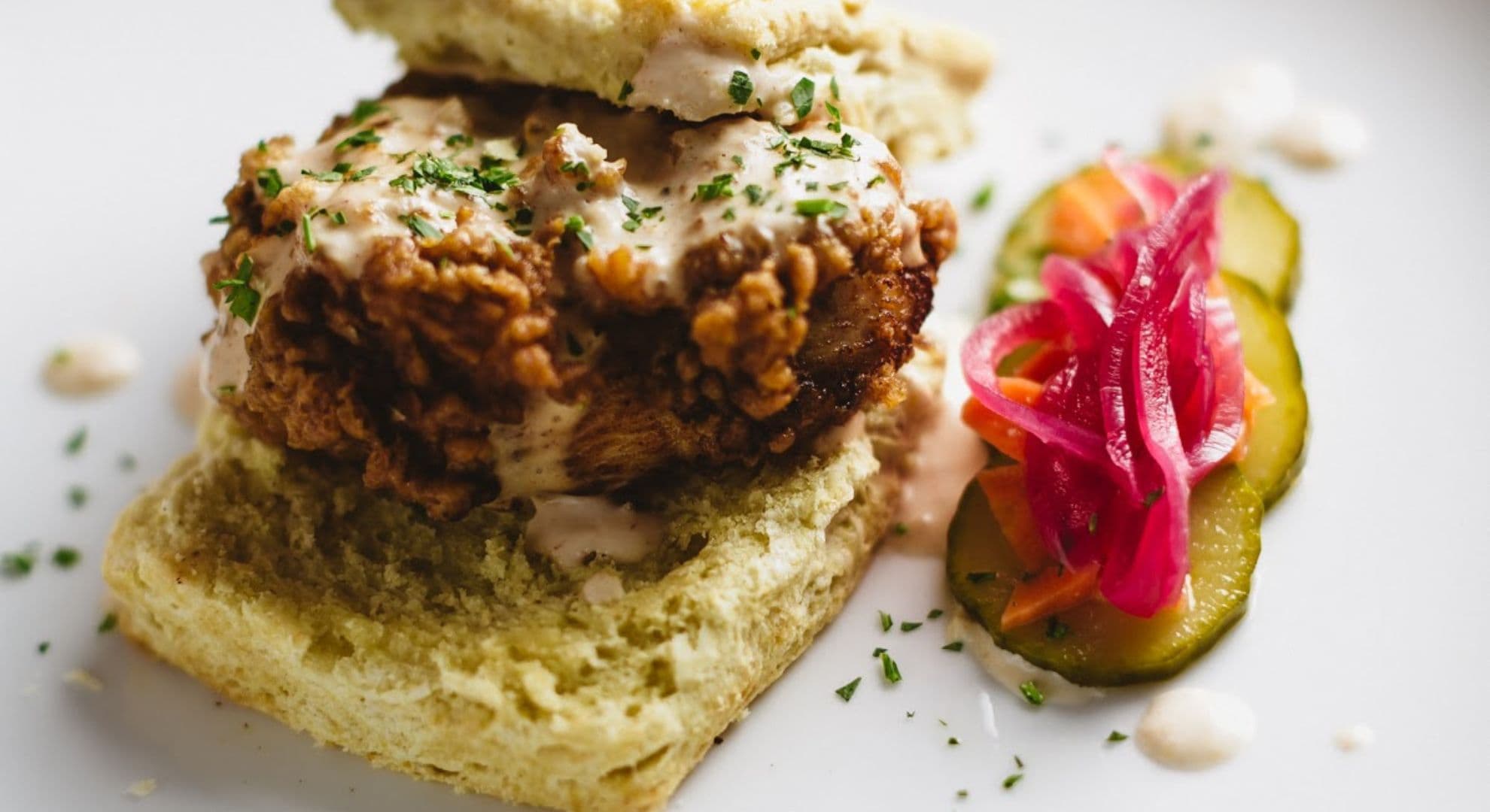 A close-up of a fried chicken sandwich topped with sauce and herbs, served alongside pickles and pickled vegetables.