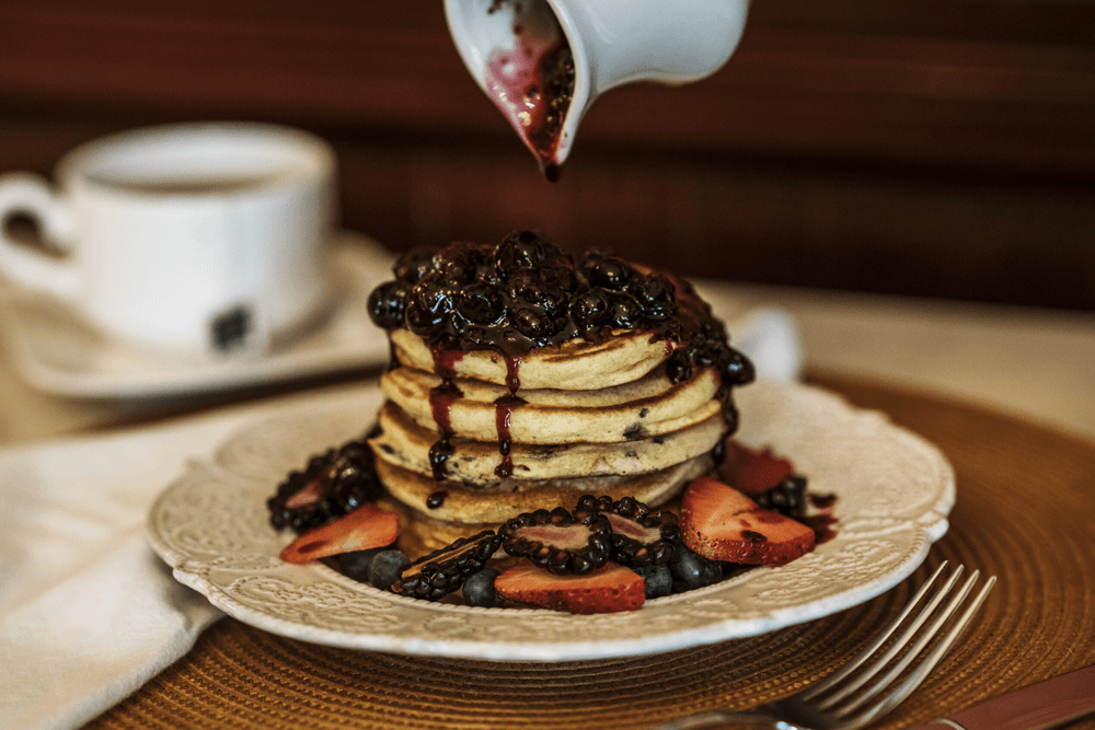 Stack of pancakes topped with berry compote, served with sliced strawberries and blueberries beside a coffee cup.