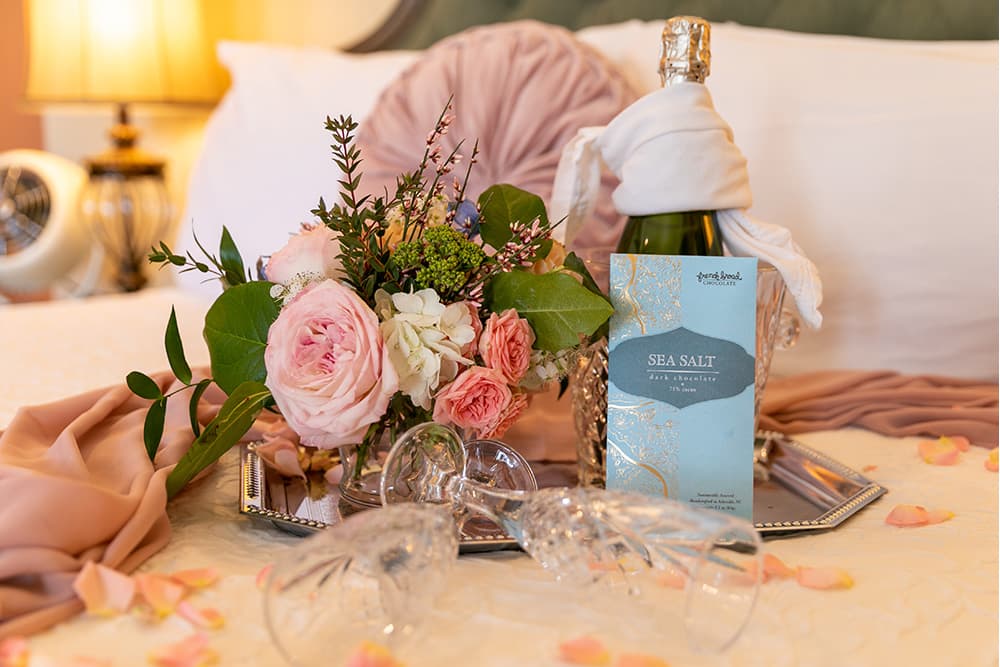 A bouquet of pastel flowers and a chocolate bar are elegantly arranged on a bed with soft fabric and rose petals.